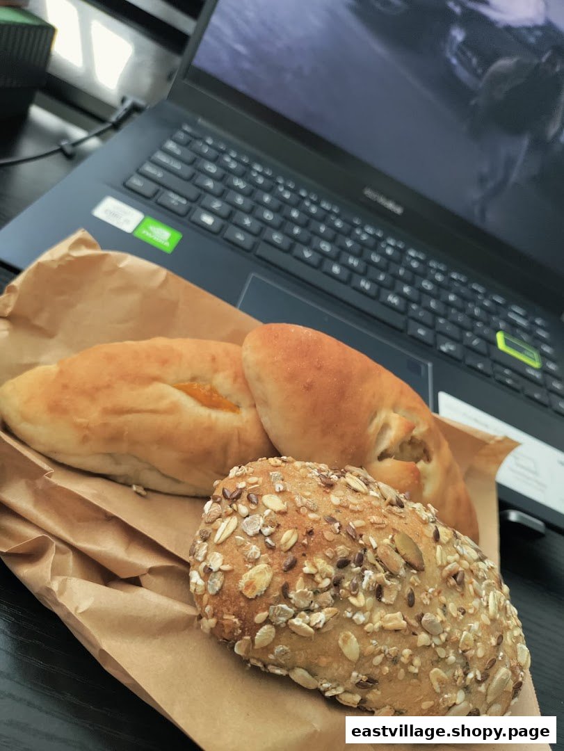 Assorted baked goods, including a multi-grain bun and filled pastries, on a paper bag next to a laptop.