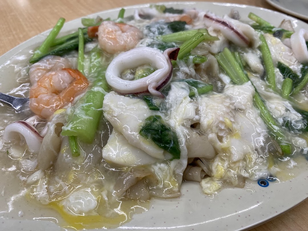 A close-up of a plate of seafood hor fun with prawns, squid, and vegetables in a rich gravy.