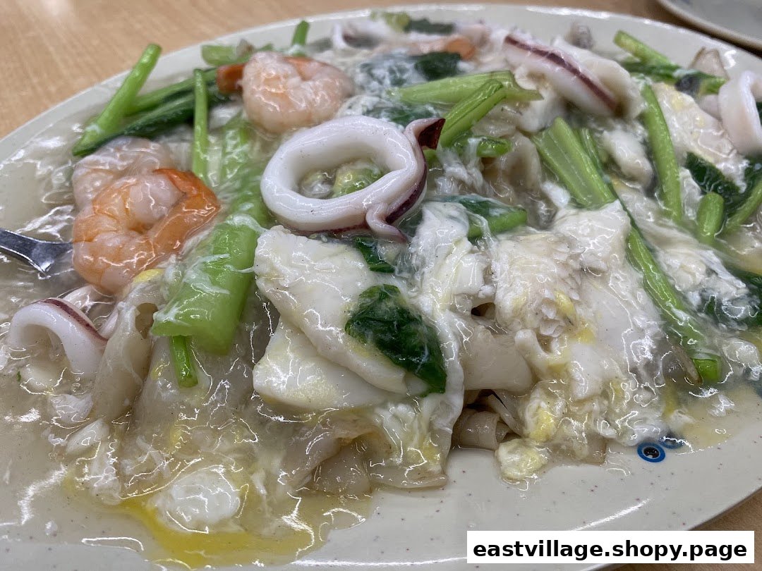 A close-up of a plate of seafood hor fun with prawns, squid, and vegetables in a rich gravy.