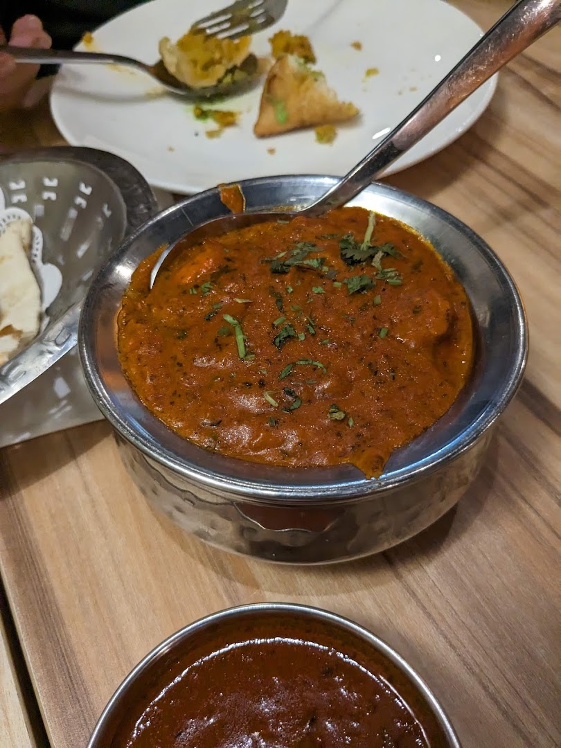 A vibrant Indian curry in a metal bowl, with a samosa and another sauce on a wooden table.