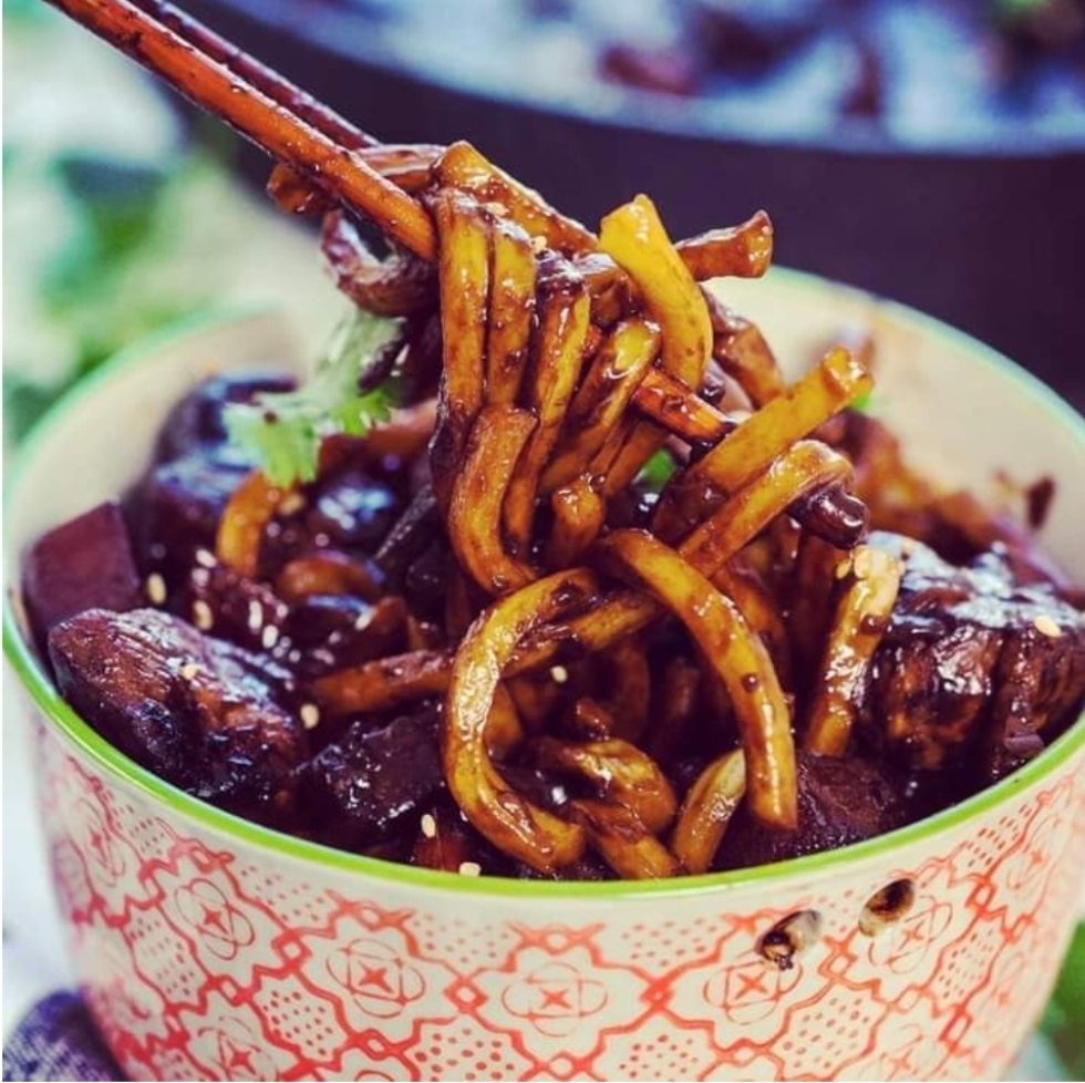 Close-up of delicious stir-fried noodles with meat and vegetables in a decorative bowl, lifted by chopsticks.