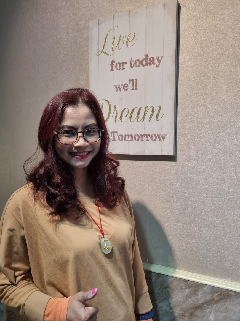 A smiling woman with red hair and glasses stands next to a motivational sign inside the salon.