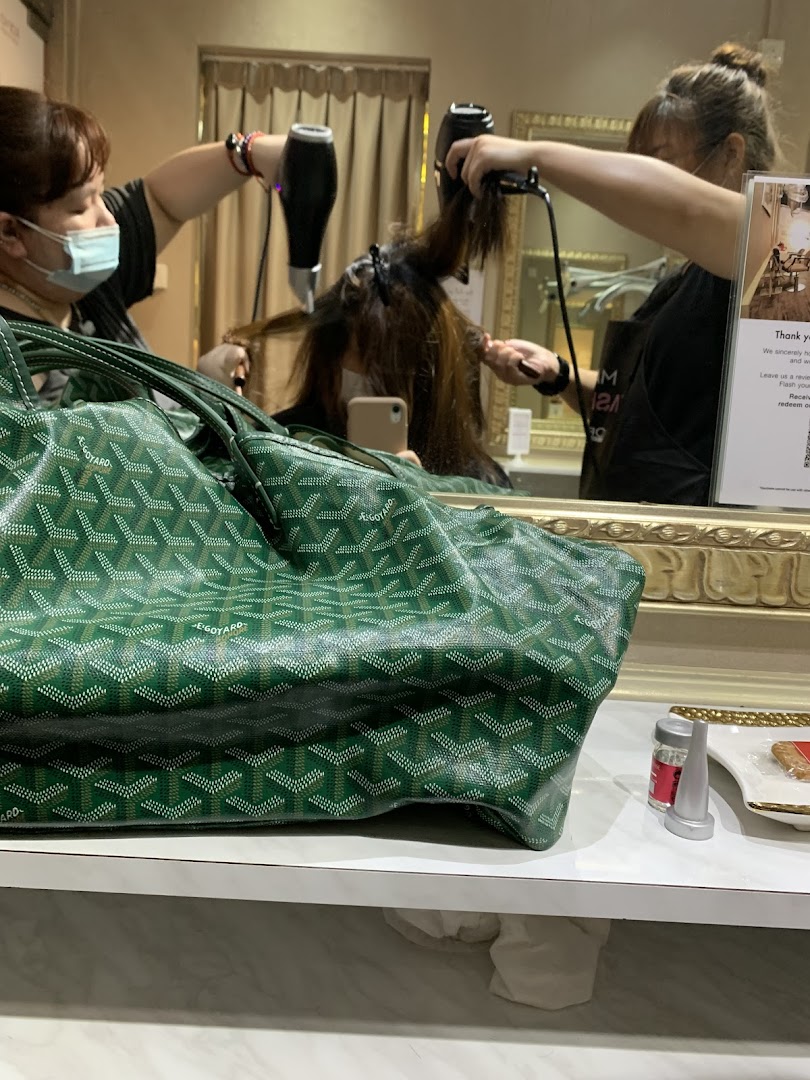 Two stylists blow-drying a client's hair in a salon, with a green bag in the foreground.