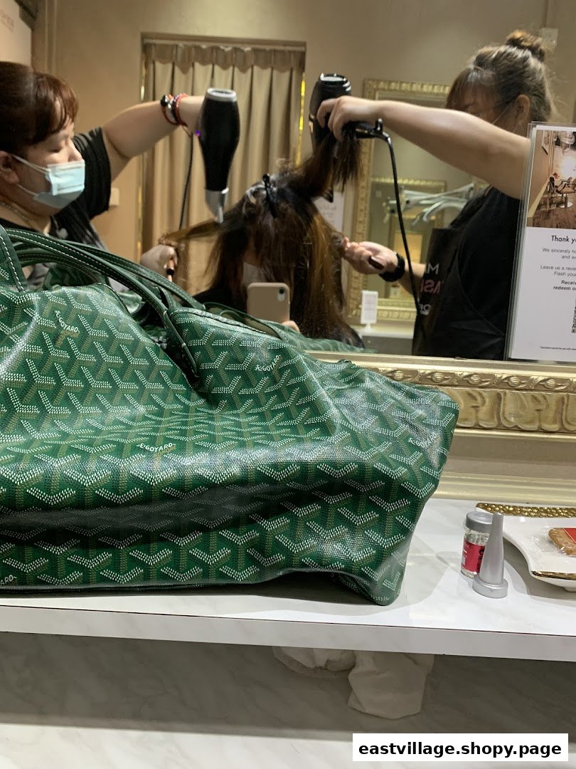 Two stylists blow-drying a client's hair in a salon, with a green bag in the foreground.