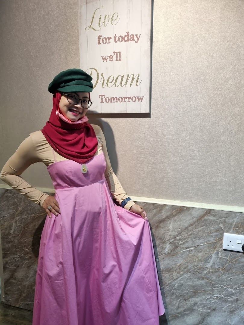 A woman in a pink dress and green hat smiles in a salon interior with a motivational sign.