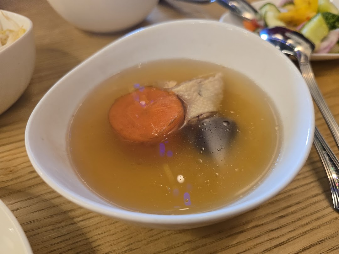 A close-up of a clear soup with carrot, meat, and mushroom in a white bowl.