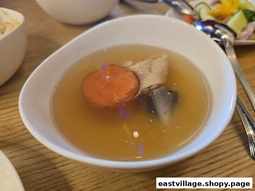 A close-up of a clear soup with carrot, meat, and mushroom in a white bowl.