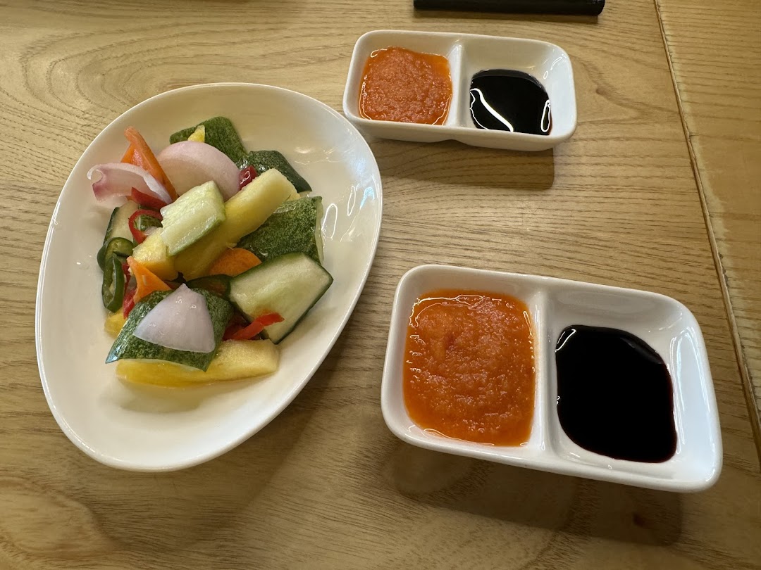 A plate of colorful pickled vegetables with two small dishes of chili and dark sauces on a wooden table.