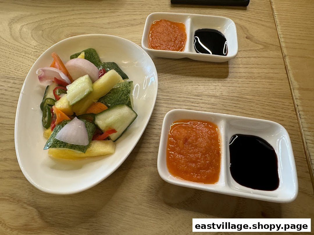 A plate of colorful pickled vegetables with two small dishes of chili and dark sauces on a wooden table.