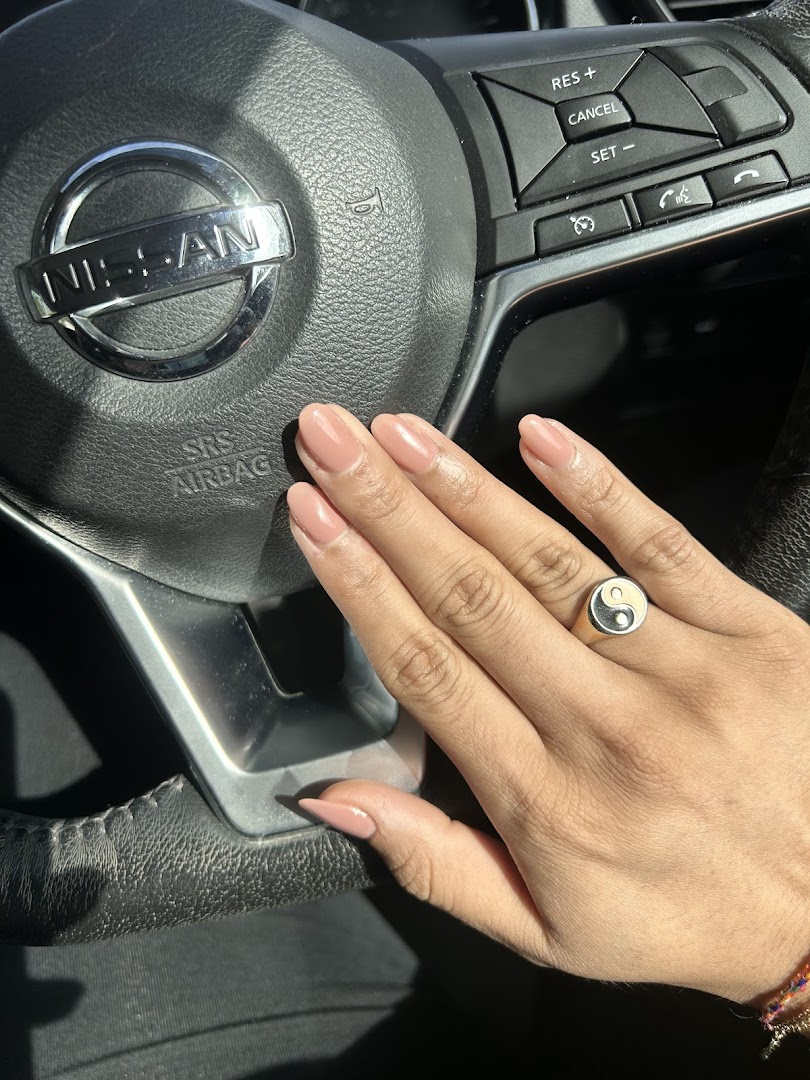 A hand with perfectly manicured nude nails and a ring, resting on a car's steering wheel.