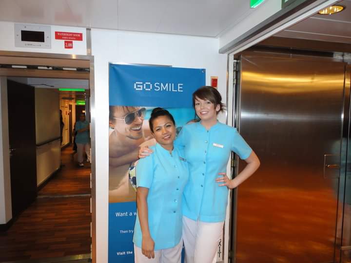 Two salon staff members in light blue uniforms stand beside a 'Go Smile' banner in an interior hallway.