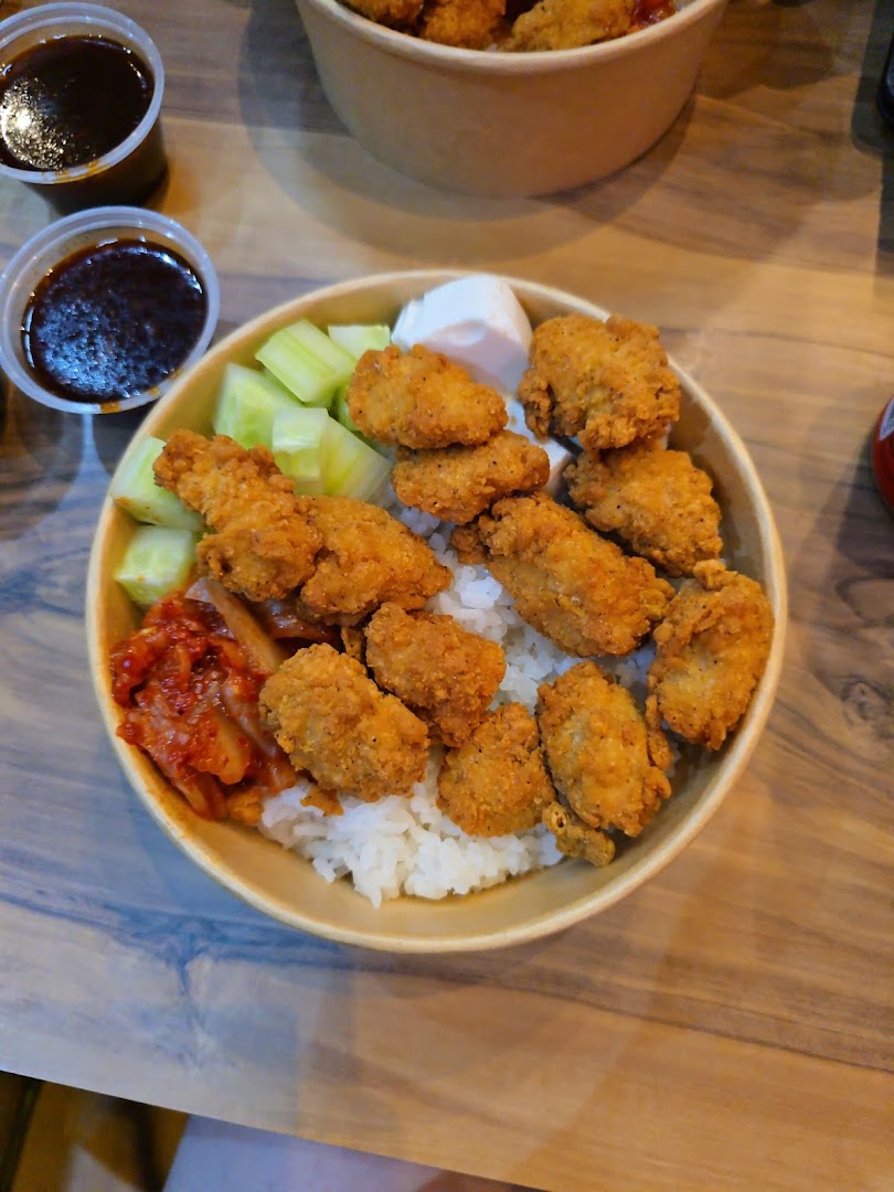 A close-up of a delicious Korean fried chicken rice bowl with kimchi, cucumber, and dipping sauces.