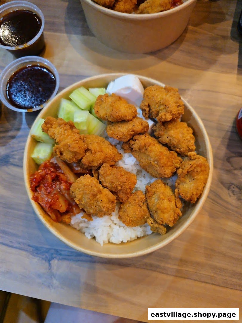 A close-up of a delicious Korean fried chicken rice bowl with kimchi, cucumber, and dipping sauces.