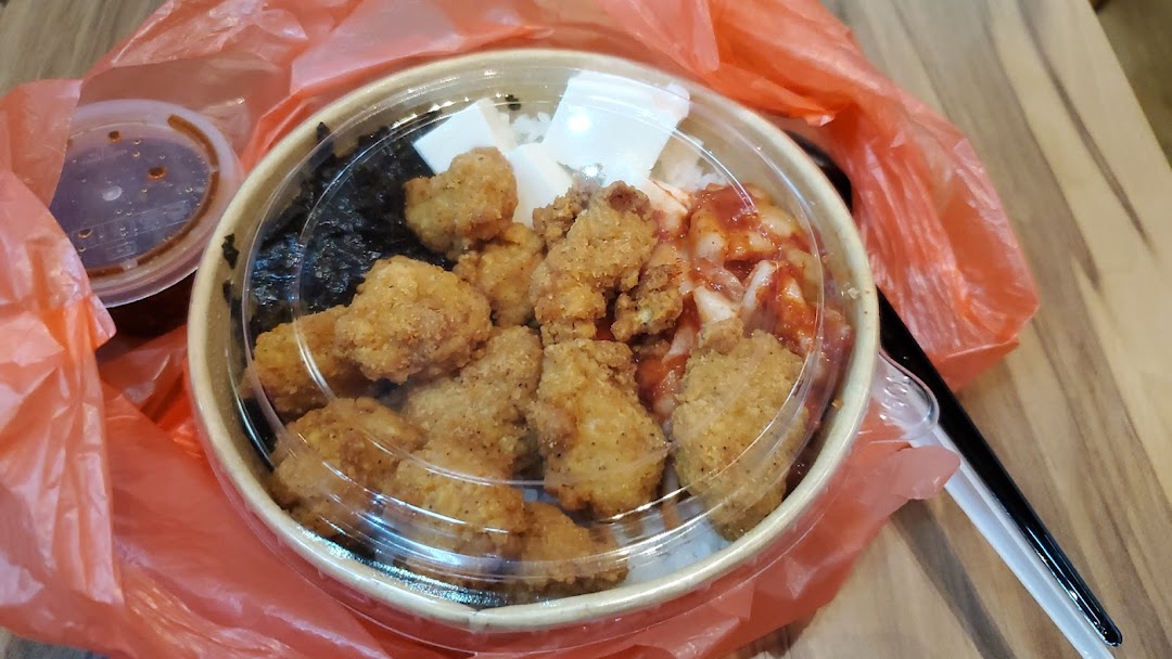 A takeaway bowl featuring Korean fried chicken, rice, kimchi, and seaweed, with a side sauce.
