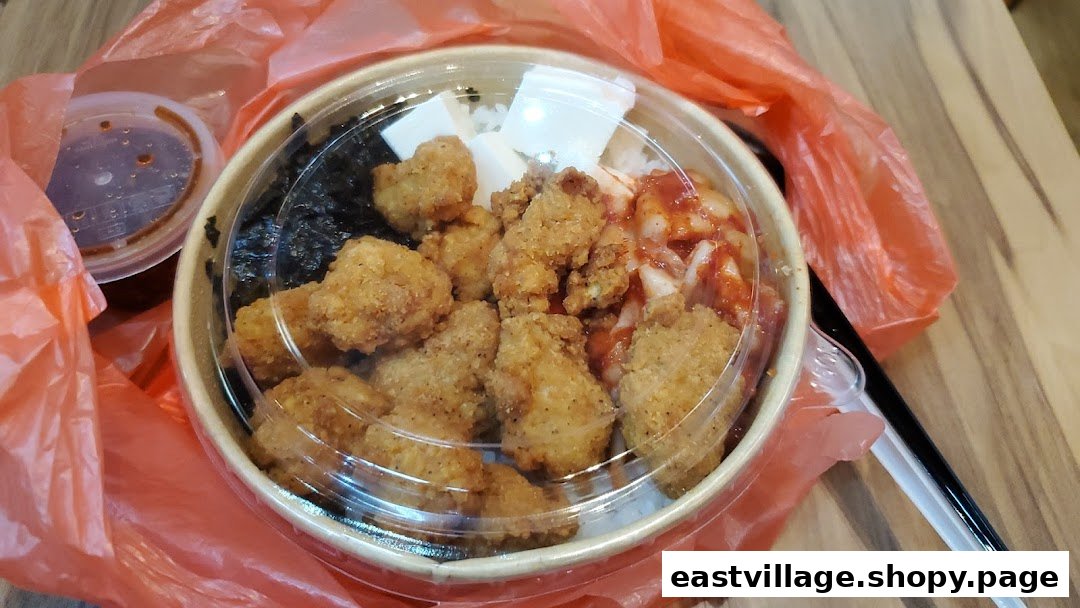 A takeaway bowl featuring Korean fried chicken, rice, kimchi, and seaweed, with a side sauce.