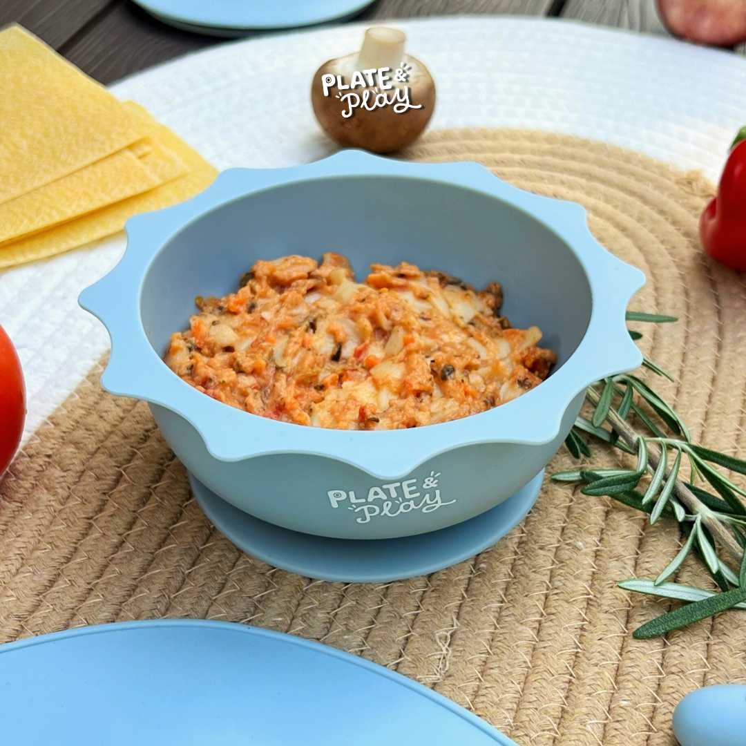 A light blue silicone bowl from Plate & Play filled with food, surrounded by fresh ingredients.
