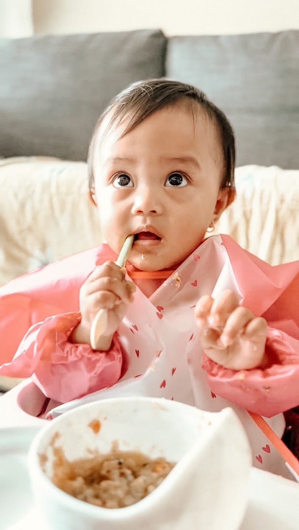 A cute baby in a high chair, wearing a pink bib, eating from a bowl with a spoon.