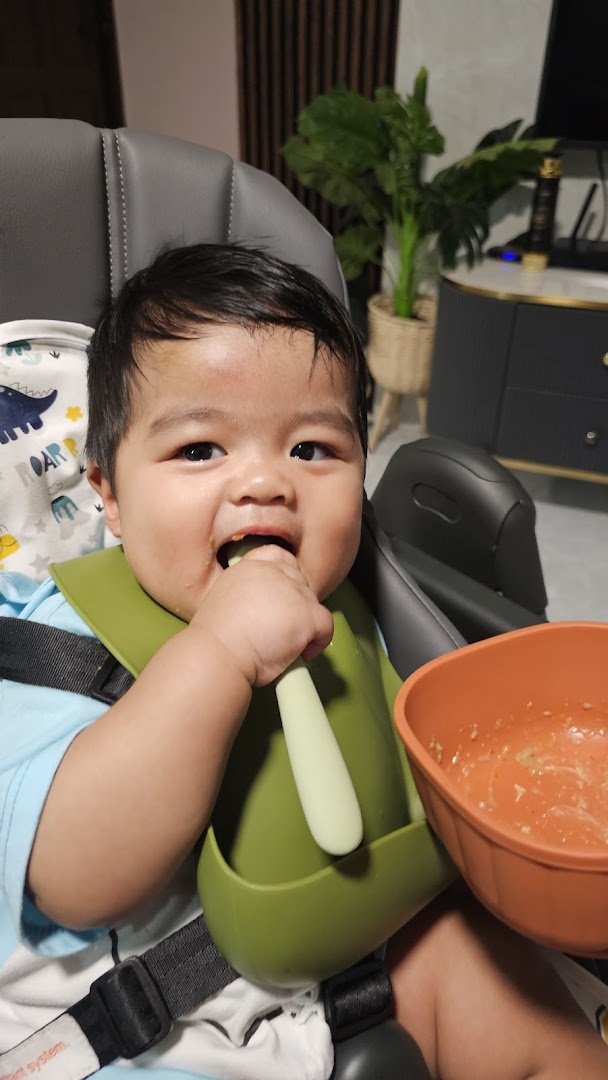 A happy baby in a high chair, wearing a bib and eating from a bowl with a spoon.