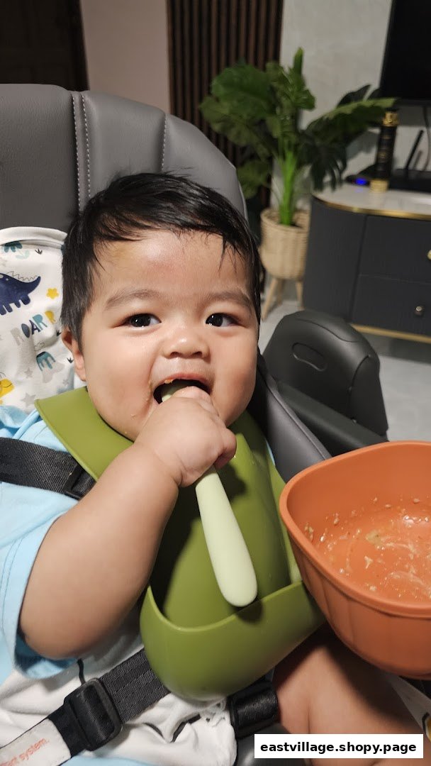 A happy baby in a high chair, wearing a bib and eating from a bowl with a spoon.
