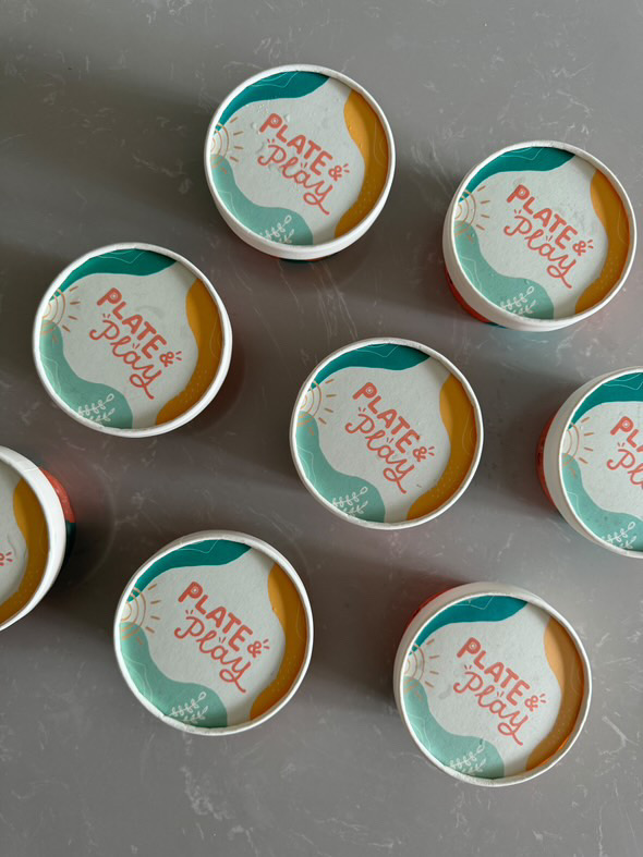 A top-down view of several colorful 'Plate & Play' branded containers on a grey surface.