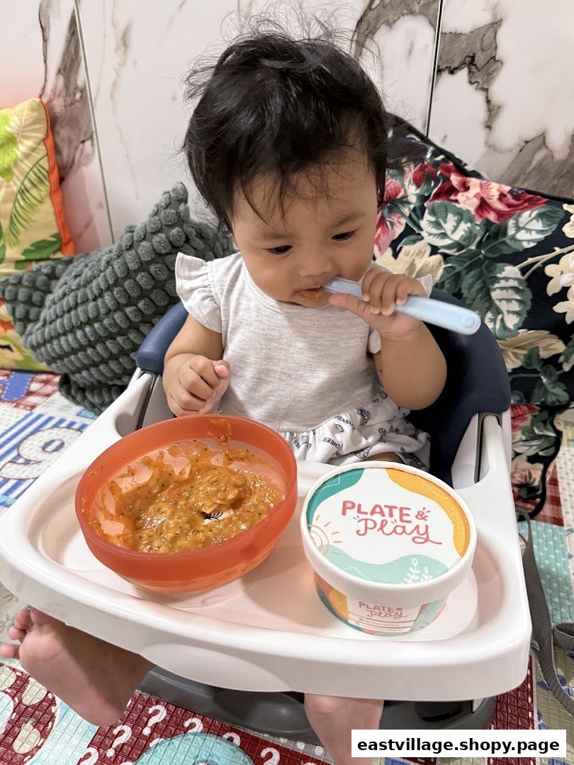 A baby in a high chair enjoying a meal from a bowl, with a 'Plate & Play' container visible.