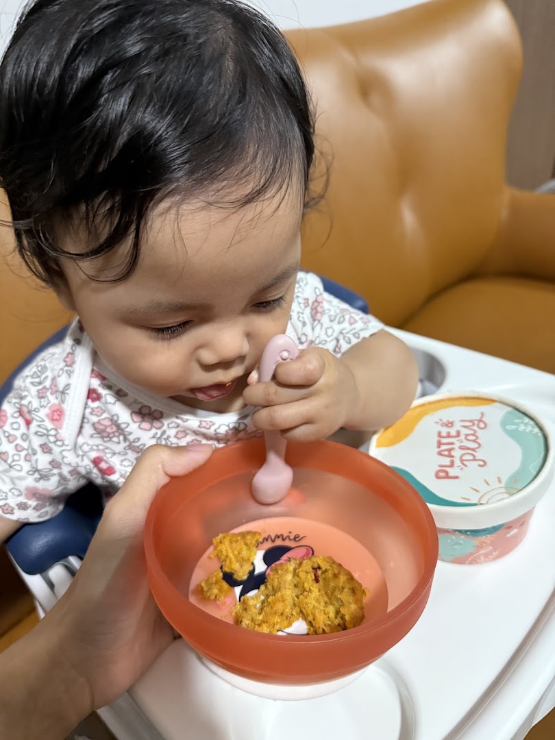 A baby in a high chair learning to self-feed with a Plate & Play bowl and spoon.