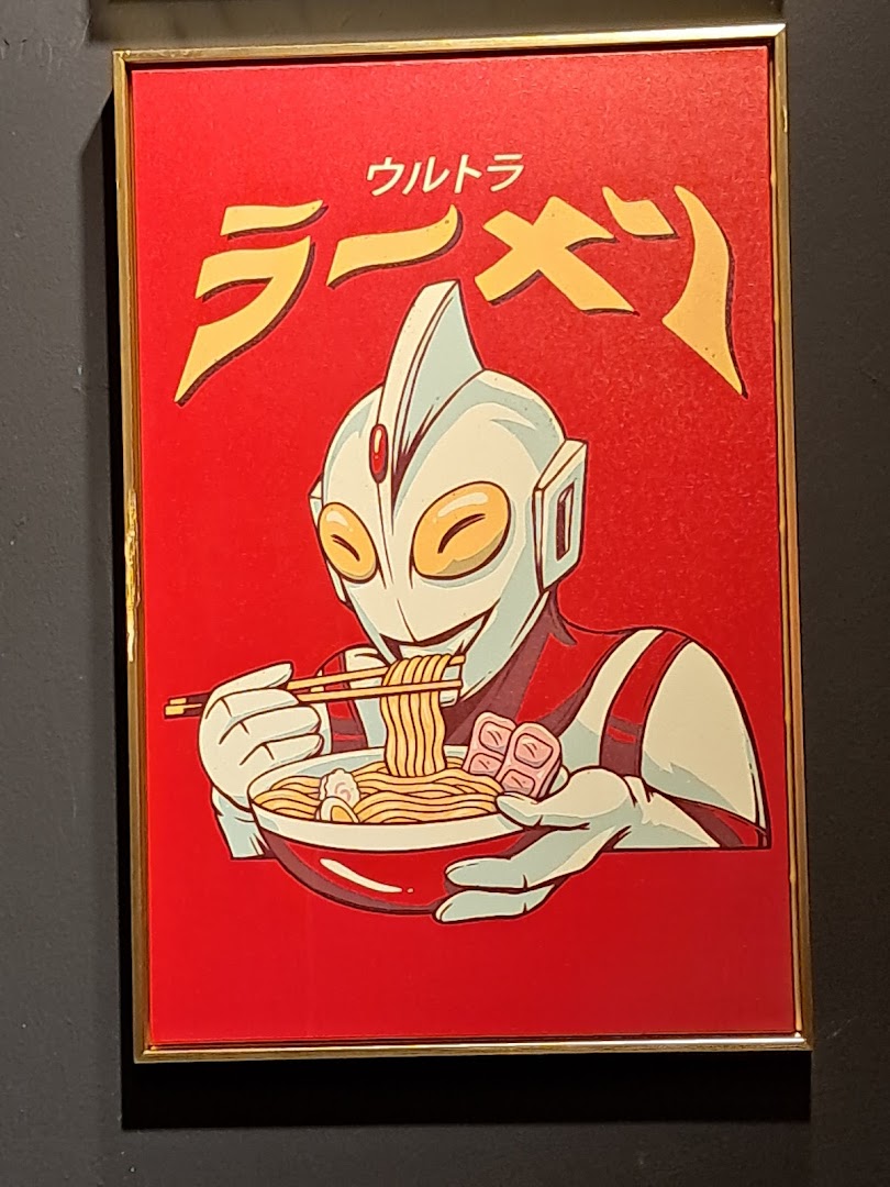 A framed poster featuring an Ultraman-like character happily eating a bowl of ramen noodles.
