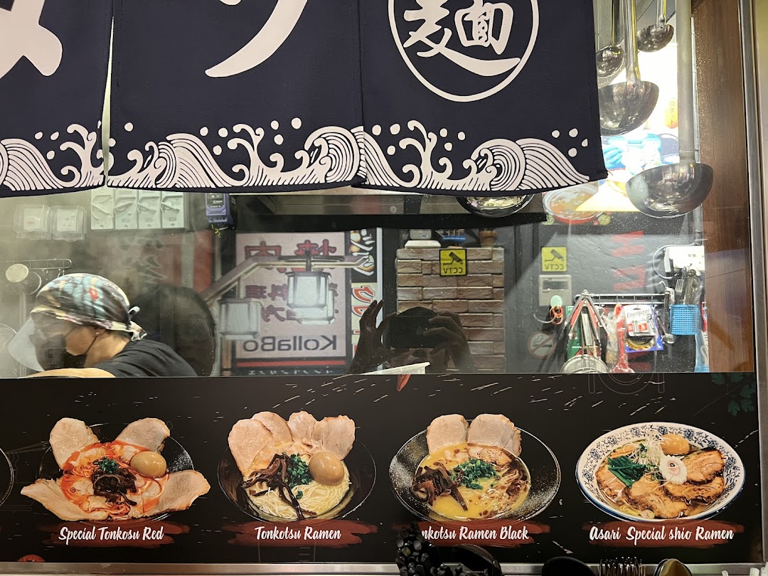A ramen shop's front displaying various ramen dishes, with a chef preparing food inside.
