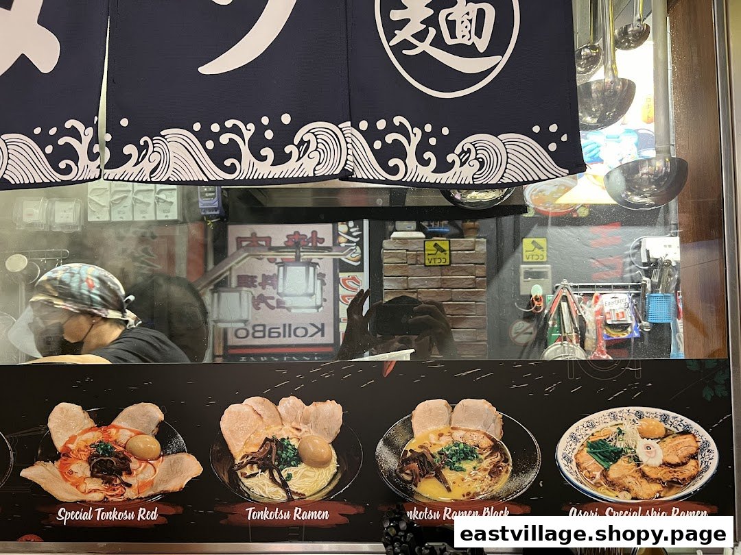 A ramen shop's front displaying various ramen dishes, with a chef preparing food inside.