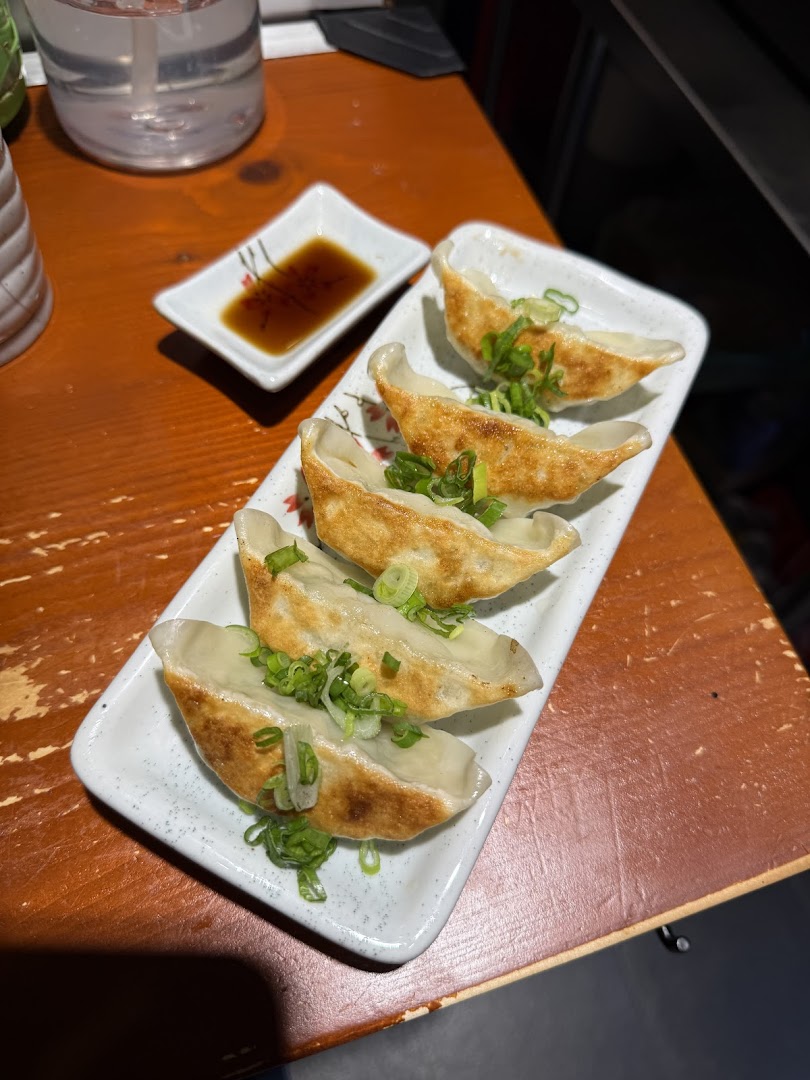 A plate of golden-brown gyoza topped with green onions, served with soy sauce on a wooden table.