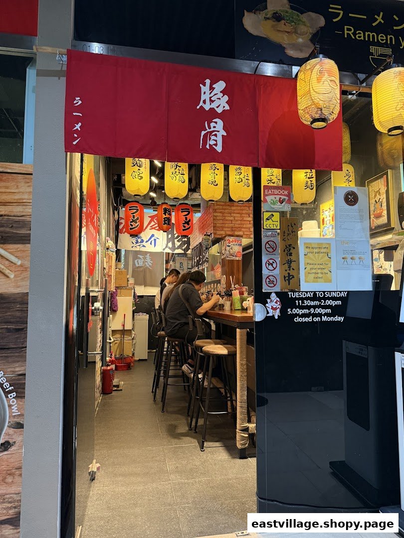 The entrance of Ramen-ya, a Japanese ramen shop, featuring a red banner, lanterns, and customers enjoying meals inside.