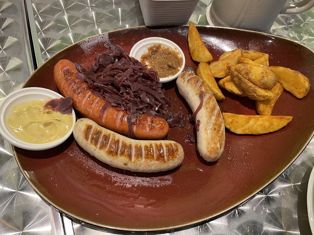 A hearty plate featuring two grilled sausages, red cabbage, potato wedges, and dipping sauces.