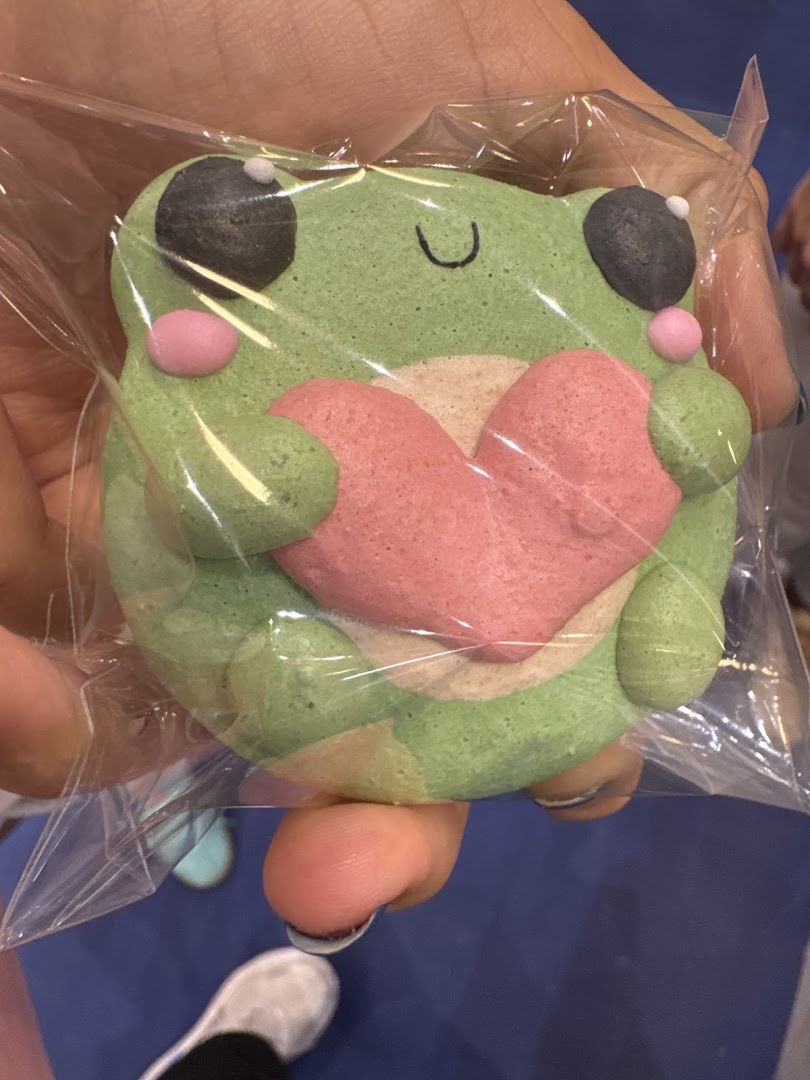 A hand holds a cute, green frog-shaped macaron with a pink heart, individually wrapped.