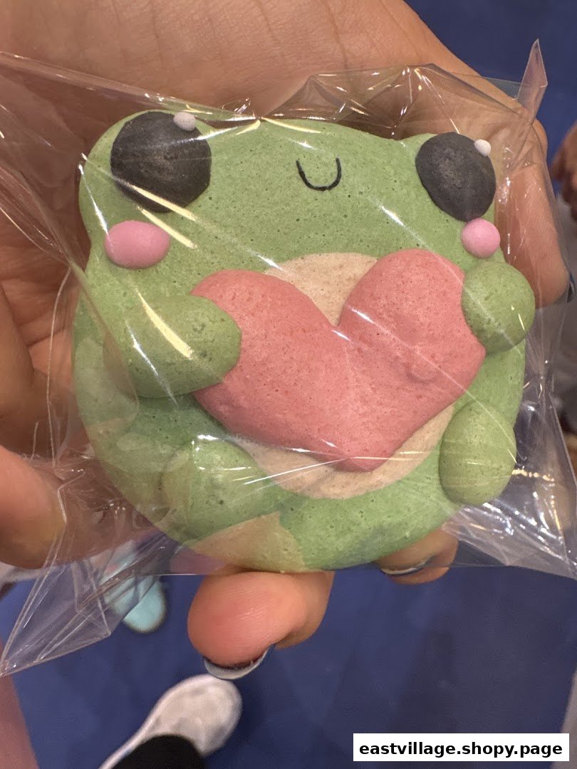 A hand holds a cute, green frog-shaped macaron with a pink heart, individually wrapped.