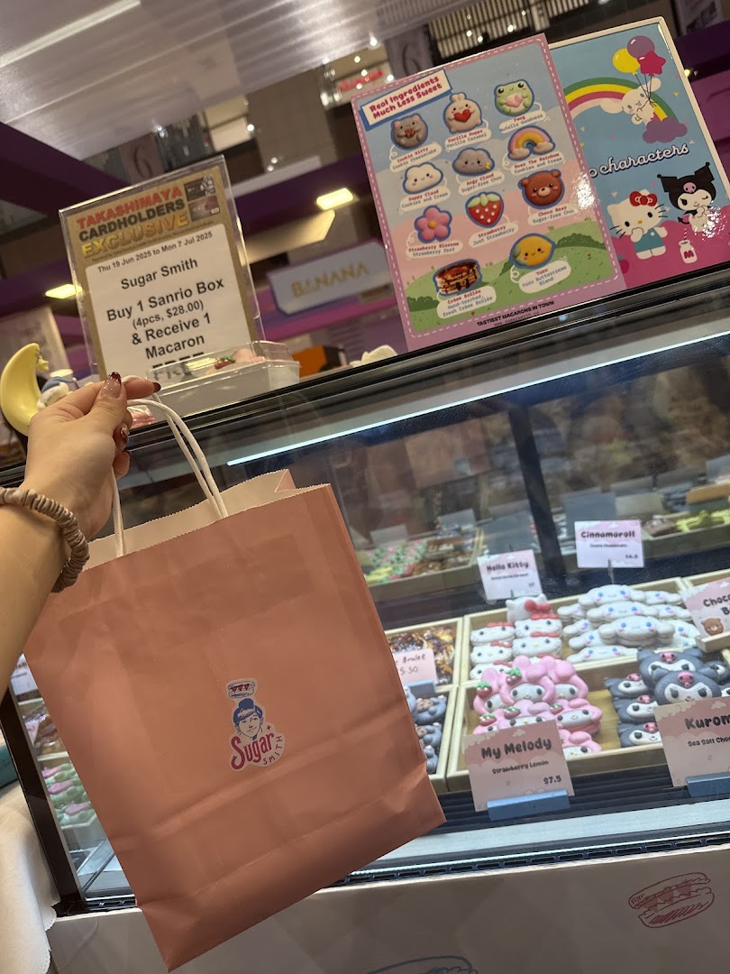 A hand holds a Sugarsmith shopping bag in front of a display case filled with adorable character macarons and promotional signs.