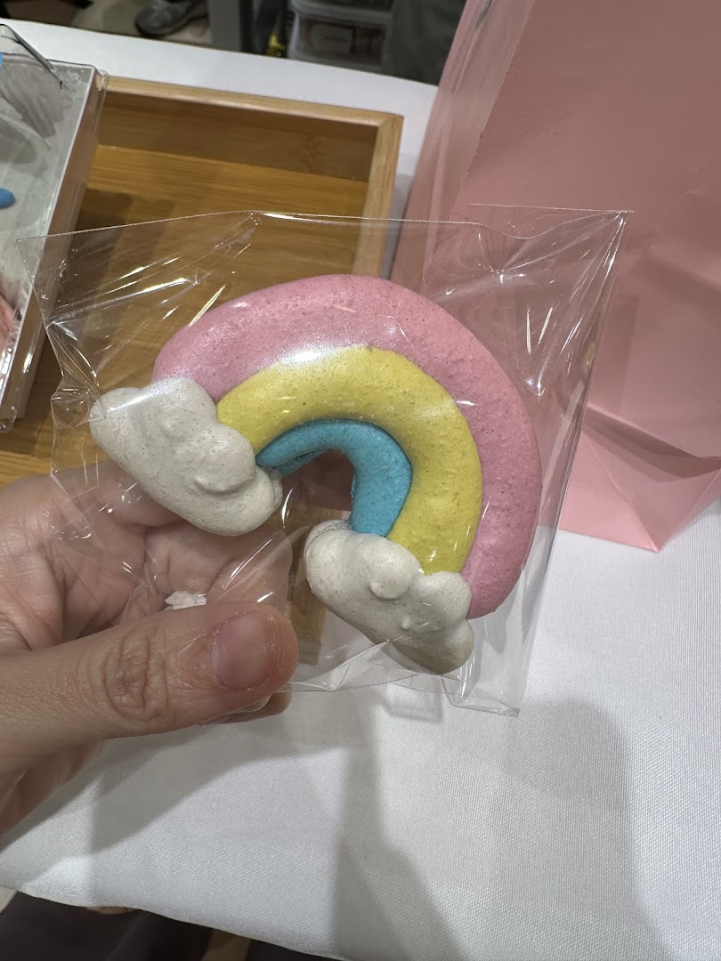 A hand holds a colorful rainbow-shaped sweet treat, likely a macaron, from Sugarsmith.