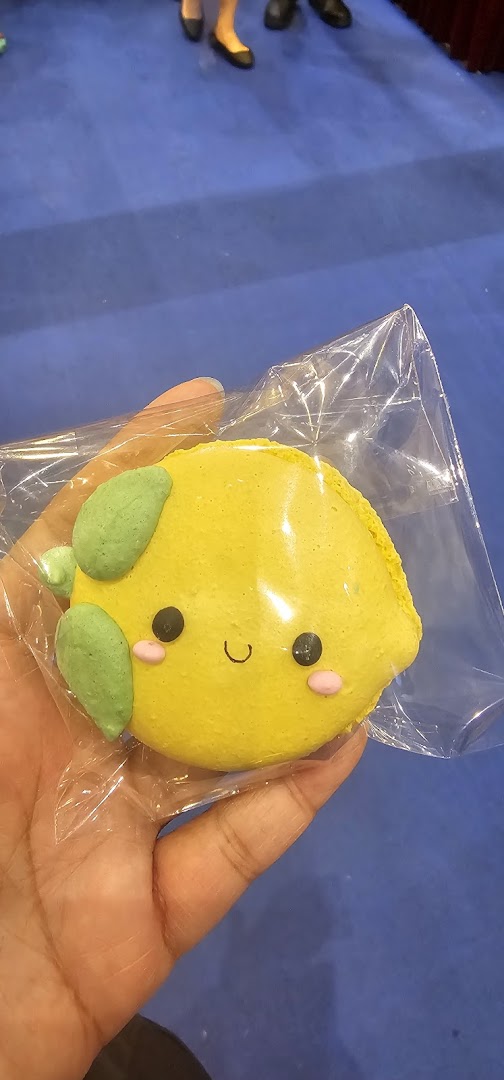 A hand holds a cute, lemon-shaped macaron with a smiling face in clear packaging.