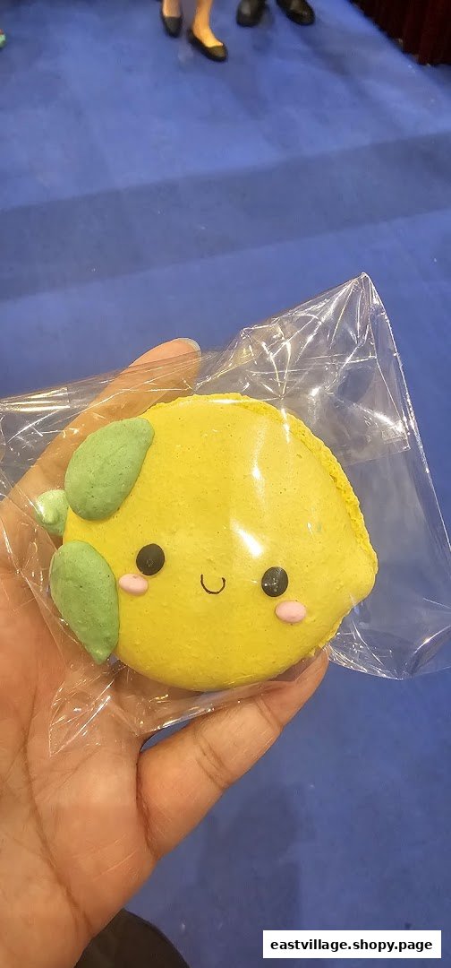 A hand holds a cute, lemon-shaped macaron with a smiling face in clear packaging.