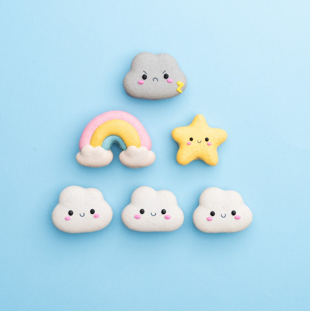 Adorable cloud, rainbow, and star-shaped cookies/macarons arranged on a light blue background.