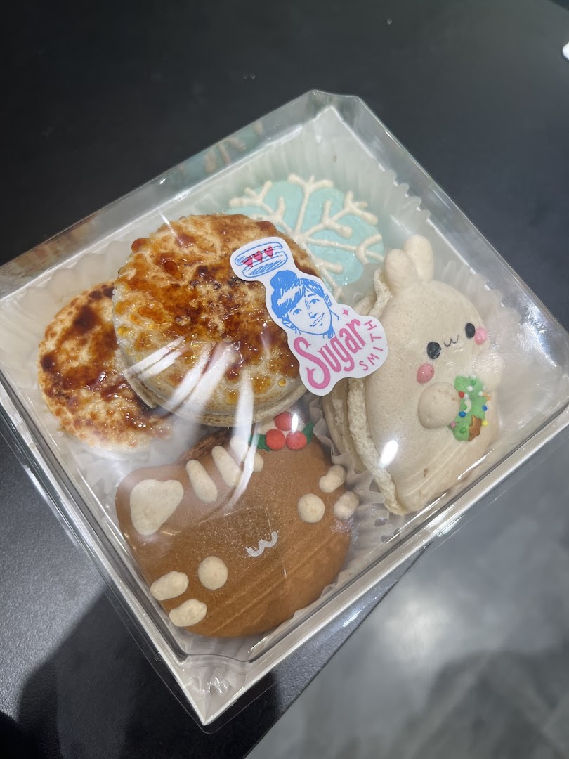 A clear box filled with assorted festive and whimsical baked goods from Sugarsmith, including cookies and tarts.