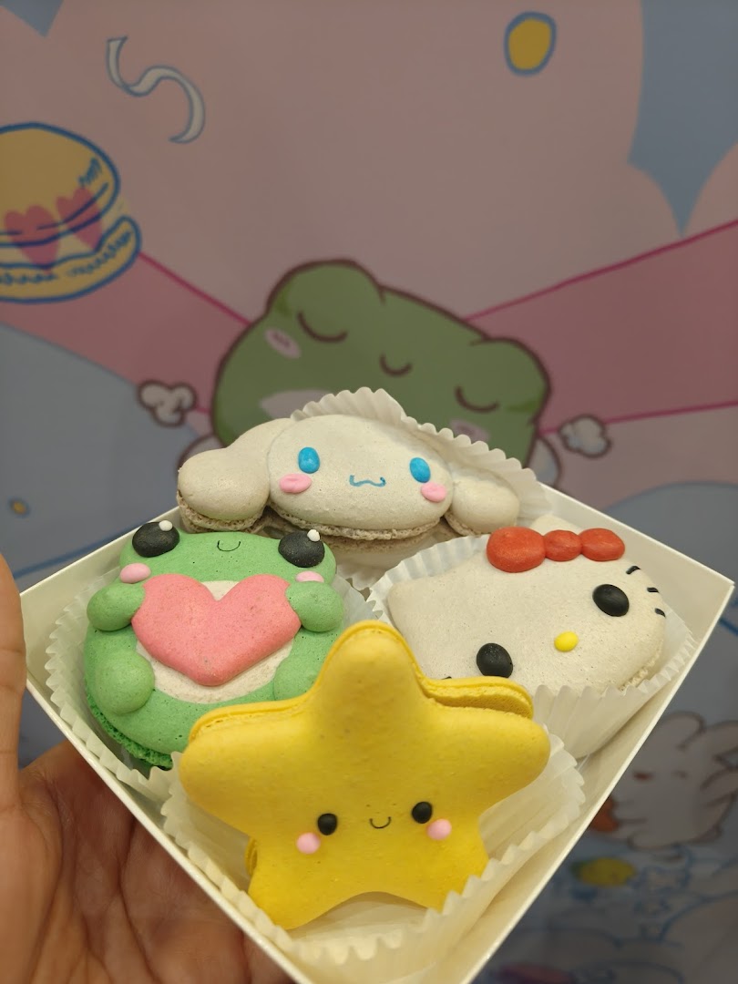 A hand holds a box of four adorable character-themed macarons, including Cinnamoroll, Hello Kitty, a frog, and a star.