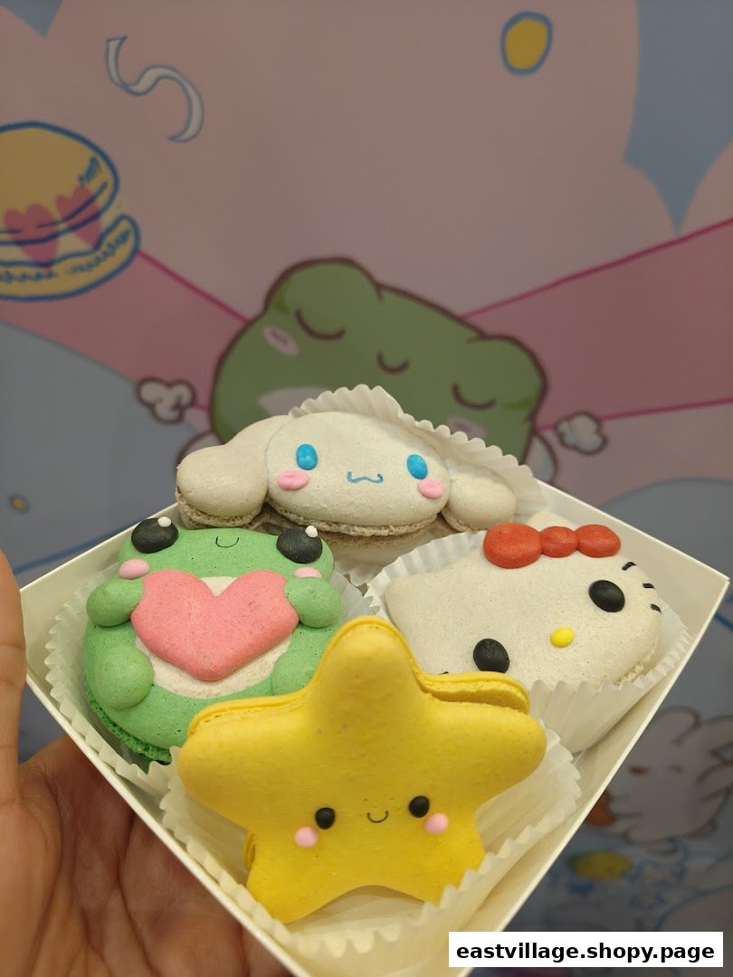 A hand holds a box of four adorable character-themed macarons, including Cinnamoroll, Hello Kitty, a frog, and a star.