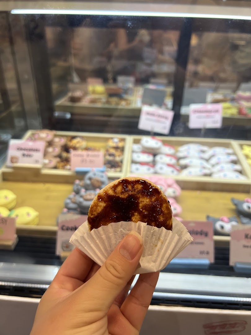 A hand holds a pastry in a paper cup, with a display case of various sweet treats behind it.