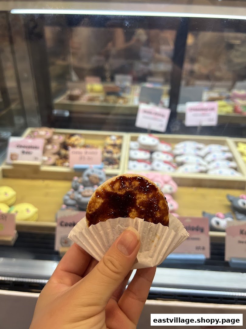 A hand holds a pastry in a paper cup, with a display case of various sweet treats behind it.
