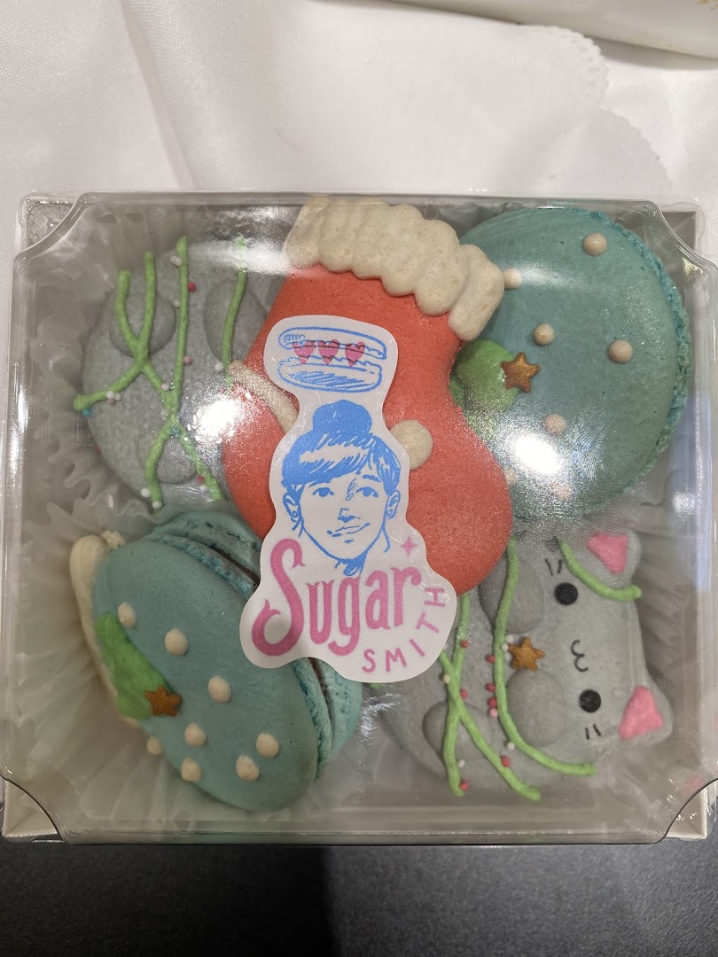 A clear plastic box filled with festive, holiday-themed macarons from Sugarsmith, featuring various shapes and decorations.