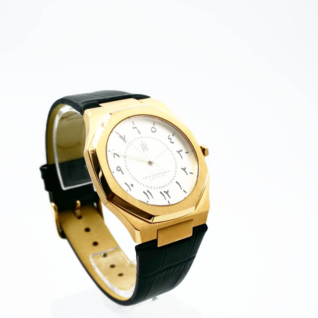A close-up of a gold-toned watch with a white dial featuring Arabic numerals and a black leather strap.