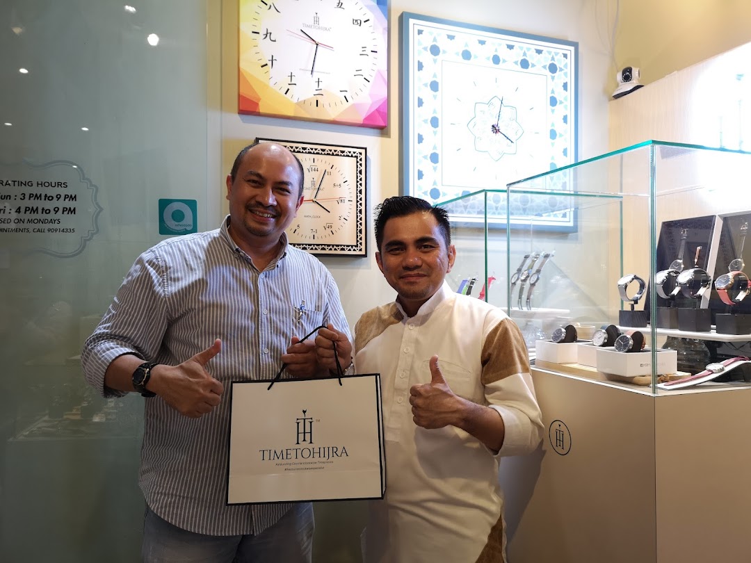 Two smiling men pose with a shopping bag inside Time To Hijra, a watch and clock shop.