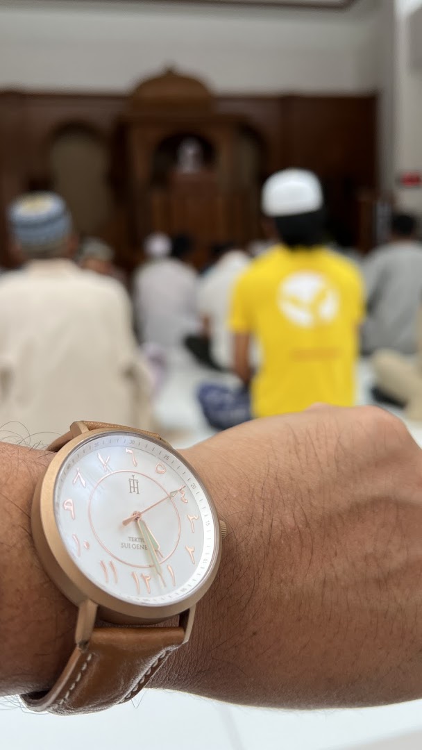 A close-up of a watch with Arabic numerals on a wrist, with blurred figures praying in the background.