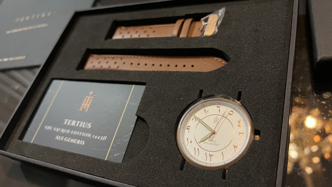 A close-up of a 'Tertius' watch with Arabic numerals and brown leather straps in its presentation box.