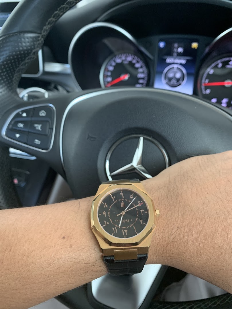 A person's arm wearing a gold watch with Arabic numerals, inside a car with a Mercedes-Benz steering wheel.